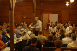 An image of people singing Sacred Harp music. It is the interior of a wooden church, where pews are arranged in a square around the leader, who stands and conducts the singing.