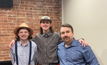 Two youthful boys wearing hats standing next to a man. The boy in the middle has his arm around the boy and man on either side of him.