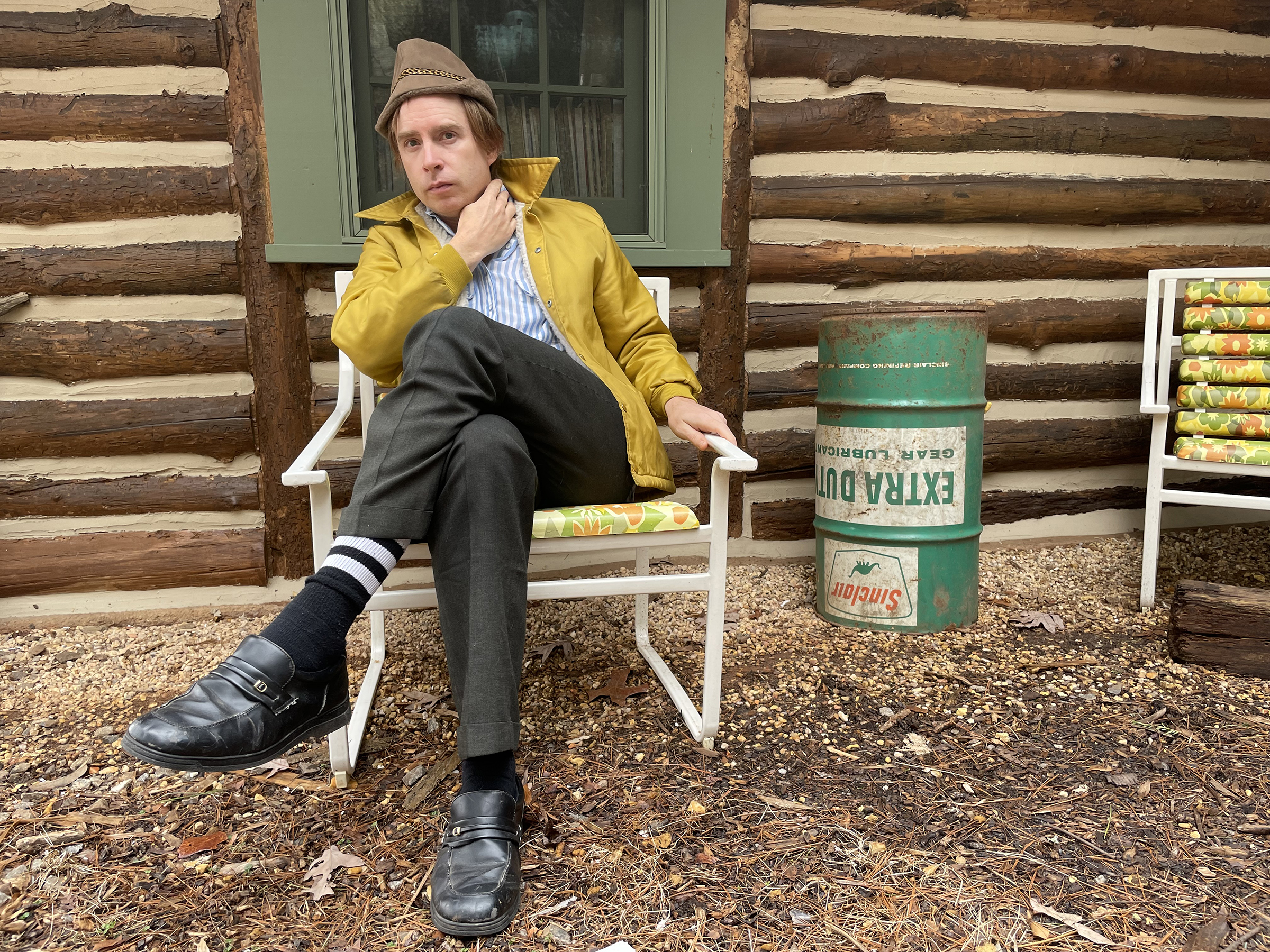 A photo of Scott McMicken sitting in front of a log cabin in a lawn chair.
