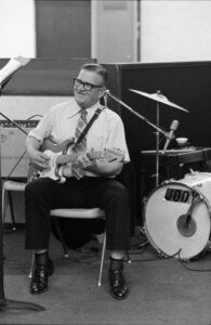 white man with glasses wearing a white short-sleeved button down shirt and tie sit in a chair playing a guitar.
