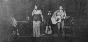 A grainy, black-and-white photo of a young Patrick Haggerty performing with Lavender Country. Only Patrick, playing guitar, and Eve Morris singing are easily visible.