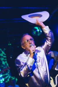 Patrick Haggerty, singing while holding up his cowboy hat. He is wearing a lavender-colored western outfit, with white tassels and accents, and holding a microphone.