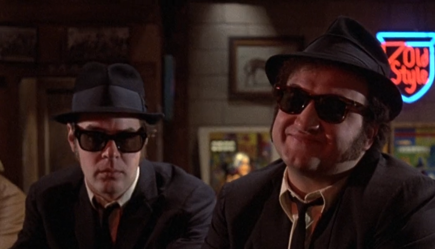 A screenshot from the scene from the 1980s film "The Blues Brothers," with the brothers making disappointed faces when told the "Country Bunker" has both kinds of music-- "Country and Western."